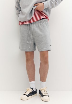 Personne portant un short de sport gris, des baskets noires avec des lacets blancs, des chaussettes blanches, un t-shirt rouge et blanc à rayures, et un sweat-shirt gris.