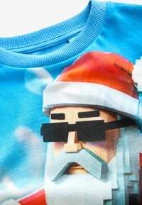 Felpa blu con un Babbo Natale in pixel art che indossa occhiali da sole e un cappello rosso, su uno sfondo azzurro chiaro con motivi di nuvole.
