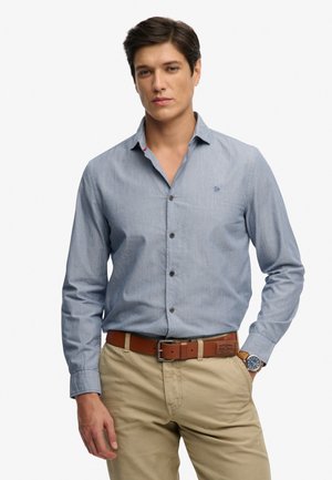 Mann trägt hellblaues Hemd, beige Hose, braunen Gürtel und Armbanduhr, steht mit einer Hand in der Tasche vor weißem Hintergrund.