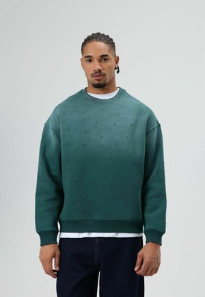 Groene trui met een ronde hals, voorzien van zwarte stippenpatronen over de stof. Zachte textuur, met geribbelde manchetten en zoom. Draagt over een wit shirt.