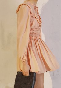 Rosa langärmliges Kleid mit Rüschelementen, tailliertem Bund und plissiertem Rock. Tragetasche aus schwarzem Textil mit Griff.