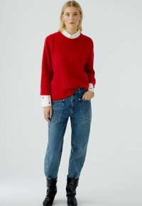 Roter Woll-Mix-Pullover mit Rundhalsausschnitt, kombiniert mit hochtaillierten blauen Denim-Jeans mit aufgesetzten Taschen und entspannter Passform, getragen mit schwarzen Stiefeln.