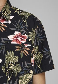 Camicia a maniche corte in tessuto nero con un vivace motivo floreale tropicale con fiori rossi e bianchi e foglie di palma verdi. Include una tasca sul petto.