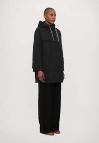 Parka noir avec capuche, cordon de serrage ajustable, deux poches avant et coupe décontractée. Fabriquée en tissu léger et texturé.