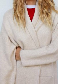 Mujer con cabello largo y rubio, vistiendo un cárdigan de punto beige sobre una blusa roja, con los brazos cruzados sobre el pecho, fondo liso y de color claro.