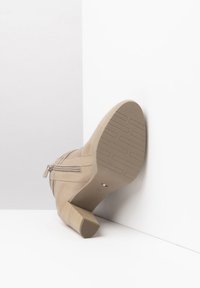 Botte de cheville beige en suède avec un talon bloc texturé. Présente une fermeture éclair sur le côté et une semelle à motif pour une meilleure adhérence.