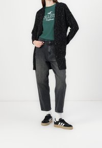 Svart stickad cardigan med en fläckig textur, bärs över en grön T-shirt, matchad med högmidjade svarta jeans och svarta sneakers med vita detaljer.