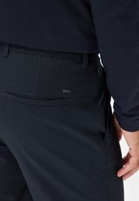 Donkergrijze broek met een gladde textuur; heeft een achterzak met een subtiele logo en een zijzak met rits. Getailleerde pasvorm.