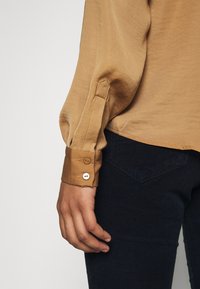 Long-sleeve blouse in beige fabric, featuring buttoned cuff details. Paired with dark blue corduroy pants with a subtle textured pattern.