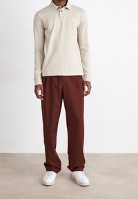 Beige langarm polo shirt met een kraag en een klein logo, gedragen met losse, donkerrode broeken en witte sneakers.