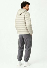 Homme en doudoune beige clair avec capuche, pantalon cargo gris, baskets blanches, debout les mains dans les poches, regardant par-dessus son épaule.