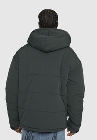 Puffertjakke i mørkegrøn med hætte, prydet med et syet, kviltet mønster. Materialet ser blødt ud med en mat finish.