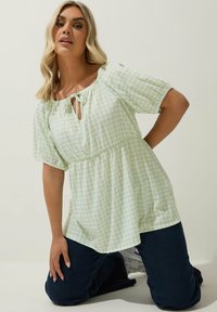 Vaaleanvihreä ruudullinen yläosa, jossa avainreikä ja solmimisnauha kaulassa, lyhyet liehuttavat hihat ja kerrostettu vyötärö, yhdistetty tummiin farkkuihin.