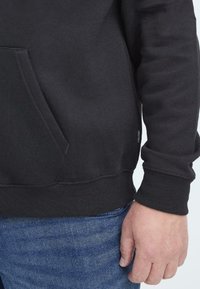 Ingrandimento del torso di una persona che indossa una felpa nera con tasca frontale e jeans in denim blu, con la mano destra rilassata lungo il fianco.