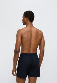 Μαύρο μαγιό βερμούδα με ελαστική μέση και πίσω τσέπη, εμφανίζεται σε αρσενικό μοντέλο από πίσω, πάνω σε λευκό φόντο.