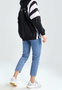 Person som bär en svart och vit huva jacka, ljusblå avklippta jeans och vita sneakers, som går med lyft vänster ben.