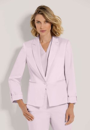 Blonde Frau mit schulterlangem Haar, die einen hellen lavendelfarbenen, maßgeschneiderten Blazer und passende Hose trägt und vor einem schlichten Hintergrund steht.