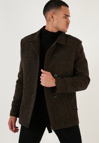 Brauner gemusterter Mantel mit doppelter Knopfleiste, schwarzen Knöpfen und Kragen. Getragen über einem schwarzen Rollkragenpullover aus texturiertem Stoff.