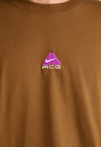 Brun bomull t-skjorte med rund hals; har en lilla og hvit brodert trekant-logo med "ACG" tekst. Glatt tekstur.