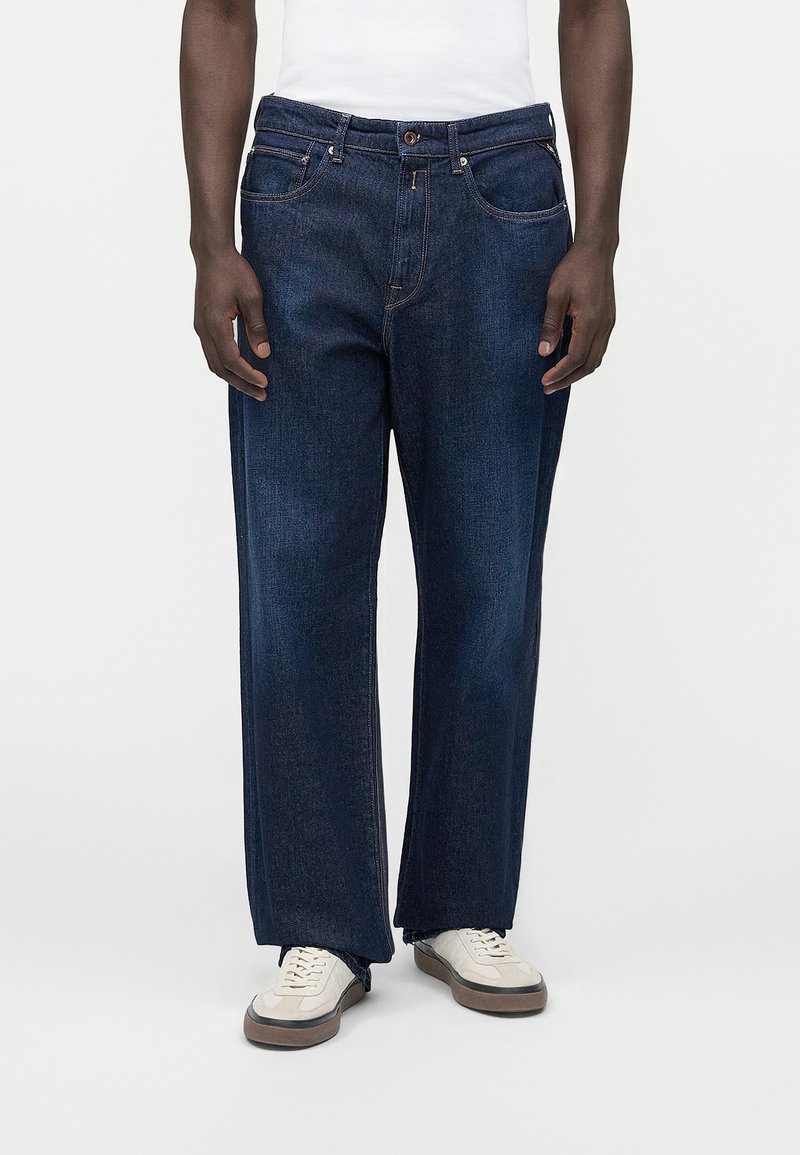 Άνδρας που φοράει σκουρόμπλε τζιν παντελόνι κανονικής γραμμής, λευκά αθλητικά παπούτσια και λευκό πουκάμισο σε απλό ανοιχτό φόντο.