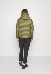 Olijfgroene pufferjas met capuchon, met een gequilt textuur, smalle pasvorm en een figuurgetailleerd ontwerp. Draagt bij zwarte broeken en sneakers.