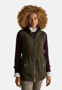 Olívovo zelená fleece vesta so zipsom vpredu, bočnými vreckami, s tunelovou šnúrkou v páse a vysokým golierom, prekrytá burgundským sveterom a vzorovanými nohavicami.