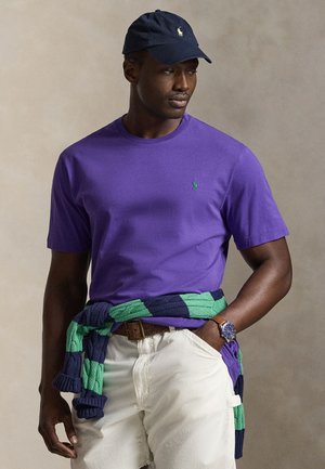 Mężczyzna w fioletowej koszuli z krótkim rękawem, białych spodniach, granatowo-zielonym swetrze związanym w talii, granatowej czapce i zegarku na rękę, patrzący na bok.