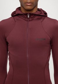 Bordová mikina na zip s vysokým límcem a přiléhavým střihem. Na hrudi je nápis "SILENCE" v černé barvě. Hladká, elastická látka.