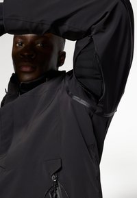 Schwarze Oberbekleidung mit strukturiertem Finish, ausgestattet mit hohem Kragen, Reißverschlüssen und belüfteten Unterarmöffnungen.
