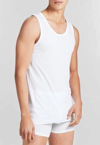 Uomo che indossa una canotta bianca senza maniche e boxer bianchi su uno sfondo neutro.