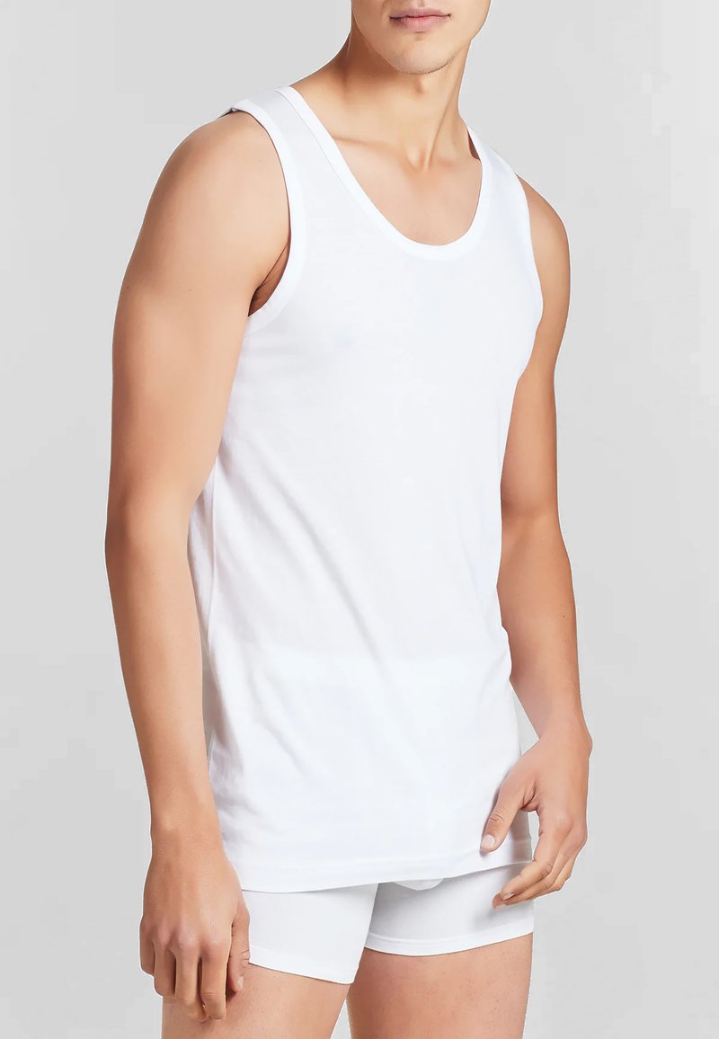 Uomo che indossa una canotta bianca senza maniche e boxer bianchi su uno sfondo neutro.