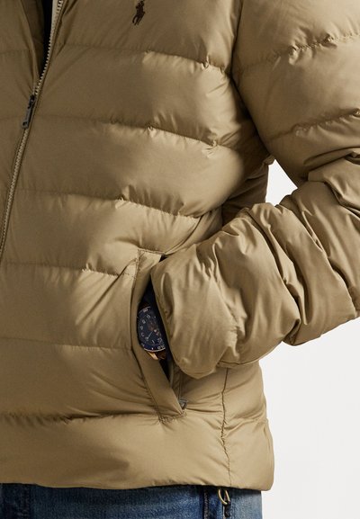 Beige puferjakke med en quiltet design, der har en frontlynlås, sidelommer og et lille logo på brystet. Glat tekstur.
