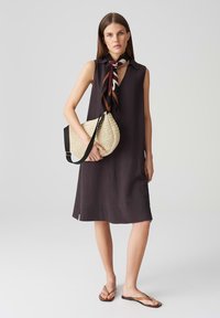 Mouwloze donkergrijze jurk met een kraag, zijsplitten en een relaxte pasvorm, gecombineerd met een strooien crossbody tas en bruine slippers.