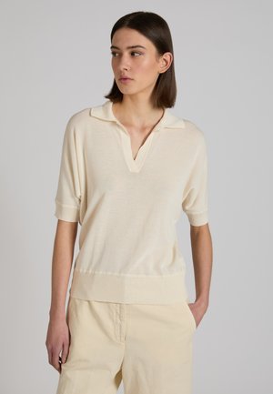 Donna con capelli castani corti che indossa un maglione beige chiaro a maniche corte con colletto e pantaloni abbinati a gamba larga, in piedi con una mano in tasca.