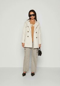 Woman wearing beige double-breasted coat, camel sweatshirt, grey cargo pants, black pointed shoes, sunglasses, holding small black handbag.