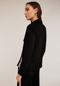 Blazer negro con un ajuste entallado, que presenta un cuello con muescas y acentos de rayas blancas a lo largo de las mangas. Textura de tela lisa.
