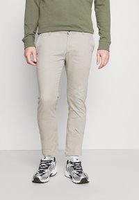 Beige chinos with a slim fit, made of cotton blend fabric. Paired with black and white running shoes. Olive green sweatshirt visible.