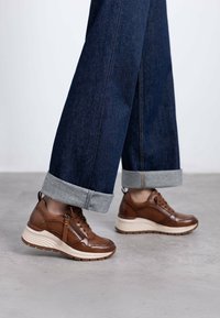 Zapatillas de cuero marrón con un acabado texturizado, con un zipper lateral y una suela intermedia de color claro, combinadas con pantalones de mezclilla oscuros ajustados en los tobillos.