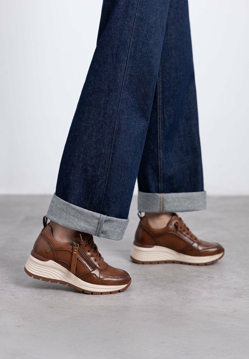 Zapatillas de cuero marrón con un acabado texturizado, con un zipper lateral y una suela intermedia de color claro, combinadas con pantalones de mezclilla oscuros ajustados en los tobillos.