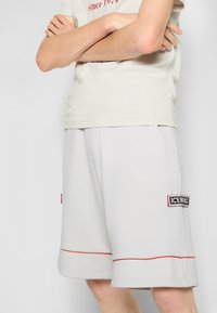 Pantaloni corti grigi chiari in felpa con accenti rossi, realizzati in tessuto morbido. Presentano tasche laterali e un patch del logo ICEBERG nero sul lato sinistro.