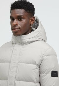 Light gray puffer jacket with a high collar and attached hood. Features quilted sections and a black fabric logo patch on one sleeve.