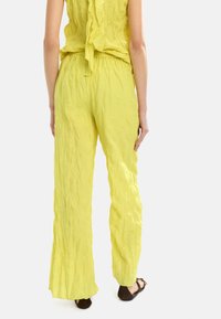 Żółte, pomarszczone spodnie z tkaniny z elastycznym pasem, szerokimi nogawkami i teksturową powierzchnią, noszone z ciemnobrązowymi płaskimi butami.