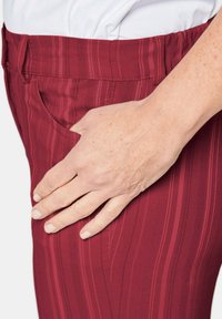 Bordeauxrode broek met subtiele verticale strepen, voorzien van een zijzak. Het materiaal lijkt zacht met een elastische tailleband. Hand rustend aan de zijkant.