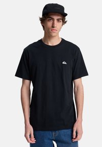 Camiseta de algodón negra con cuello redondo. Presenta un pequeño logo blanco en el lado izquierdo del pecho. Se lleva con jeans de denim azules y una gorra negra.