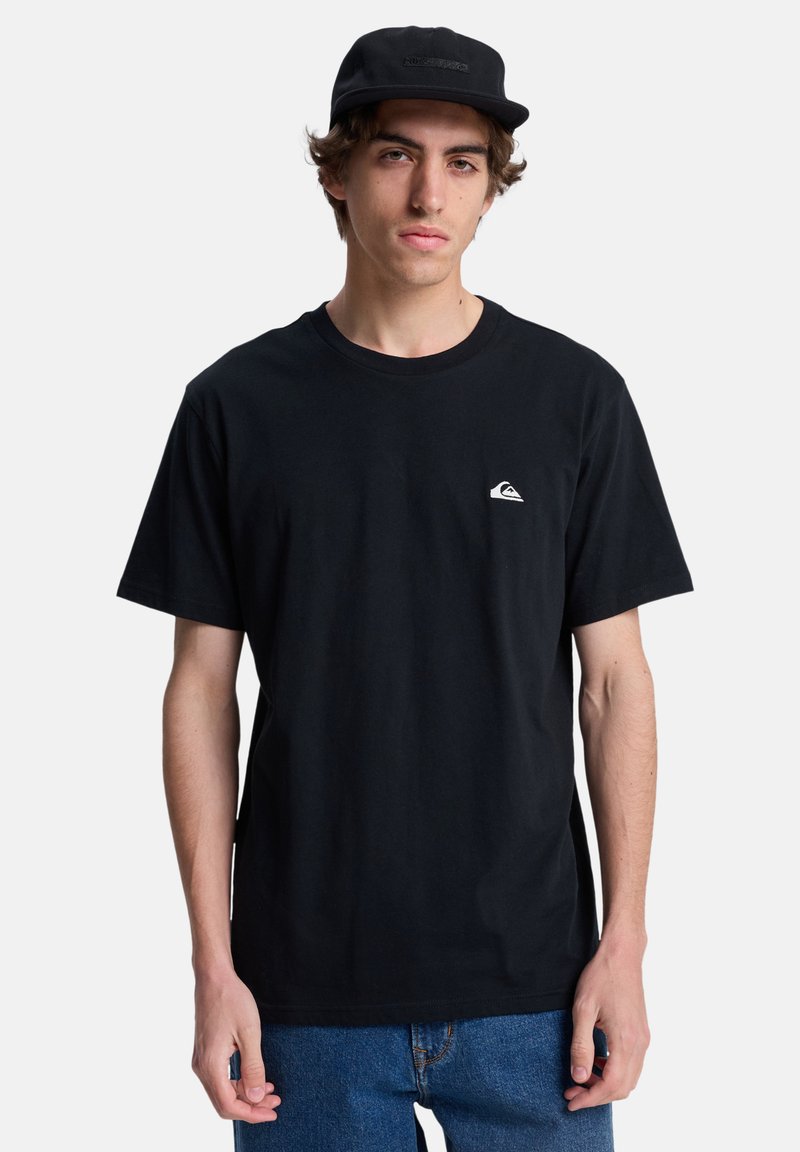Camiseta de algodón negra con cuello redondo. Presenta un pequeño logo blanco en el lado izquierdo del pecho. Se lleva con jeans de denim azules y una gorra negra.