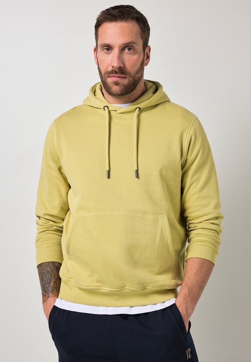 Mann mit Bart, der ein zitronengelbes Kapuzensweatshirt und marineblaue Hose trägt, steht mit den Händen in den Taschen vor einem schlichten Hintergrund.