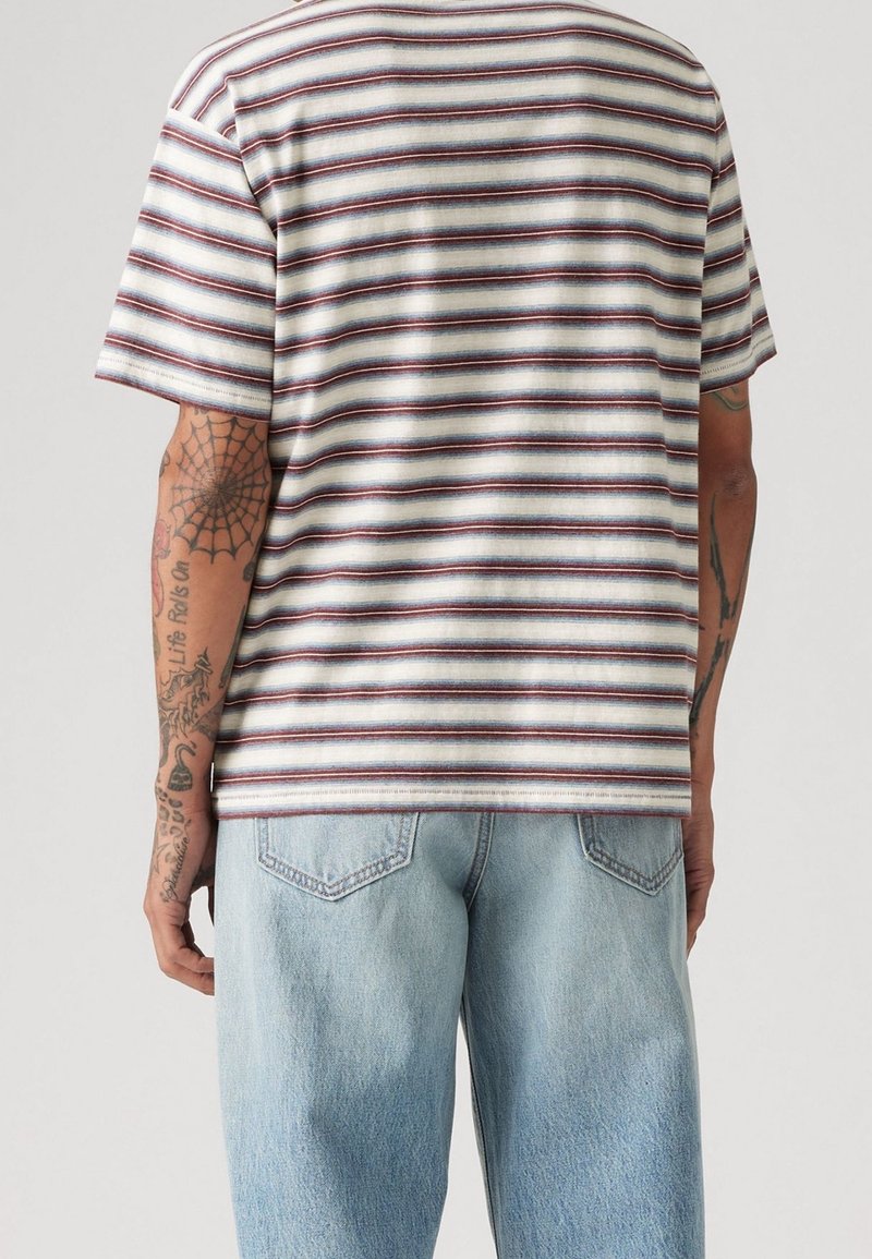 Personne portant une chemise rayée à manches courtes et un jean bleu clair, vue de dos avec des tatouages visibles sur les bras.