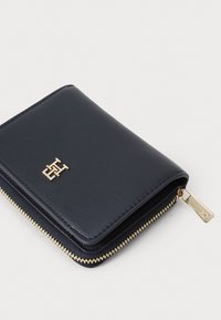 Navy leather wallet with gold zipper and hardware, featuring a subtle logo on the front and a textured finish. Compact rectangular shape.
