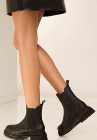 Fekete bőrből készült bokacsizma, elasztikus oldalpanelek, kerek orr és texturált talp jellemzi. A hátulján található húzófül megkönnyíti a felvételt.