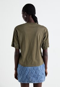 Olijfgroene katoenen T-shirt met korte mouwen, voorzien van zilveren versieringen op de mouwen, gecombineerd met een gestructureerde blauwe denim rok.